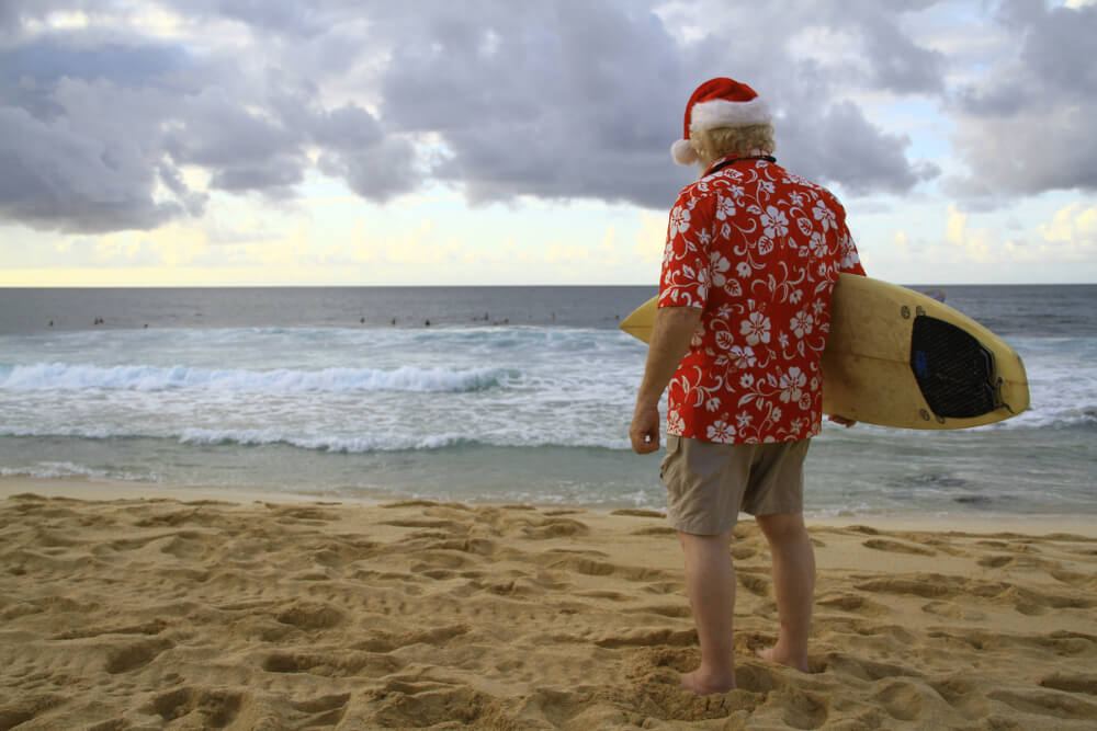 a man in a red shirt and santa hat standing on the beach with a surf board